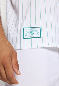 Mand hånd ved siden af hvid- og stribet Liverpool Football Club skjorte med grøn etiket og logo nær kanten.