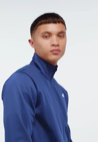 Blå zip-up jacka med hög krage, slät tyg, figursydd design och minimal branding på bröstet. Modellen poserar mot en enkel bakgrund.