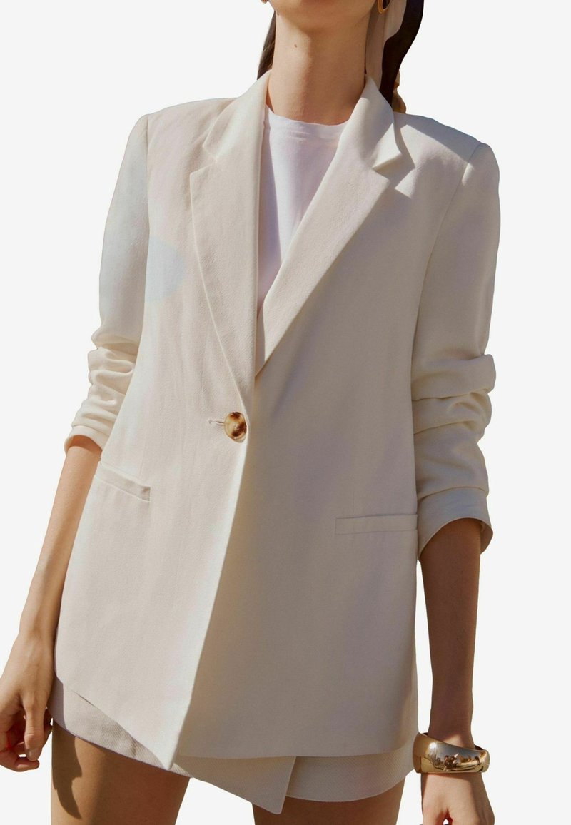 Femme portant un blazer beige clair aux manches retroussées sur un top blanc, accessoirisé d'un bracelet en or et de boucles d'oreilles créoles dorées.