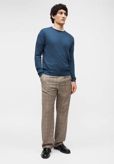 Άνδρας με σγουρά μαλλιά που φοράει μπλε πουλόβερ με στρογγυλή λαιμόκοψη, μπεζ καρό παντελόνια και μαύρα επίσημα παπούτσια, στέκεται με τα χέρια στις τσέπες.
