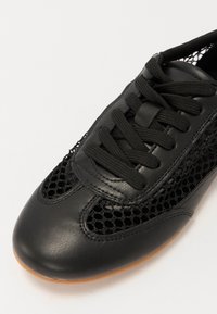 Schwarze Sneaker mit einer glatten Lederzehenkappe und Mesh-Seitenpaneelen. Runde Form mit flacher, strukturierten Gummisohle und Schnürverschluss.