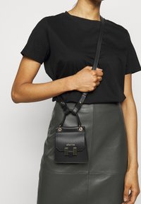 Sac à bandoulière en cuir noir avec un rabat, accents en métal doré et finition texturée. Il dispose de sangles réglables et d'un design compact.