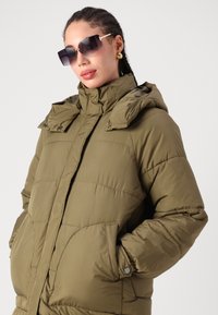 Casaco puffer verde-oliva com capuz, apresentando secções acolchoadas horizontais, fecho zip e bolsos grandes. Tecido texturizado com um acabamento suave.