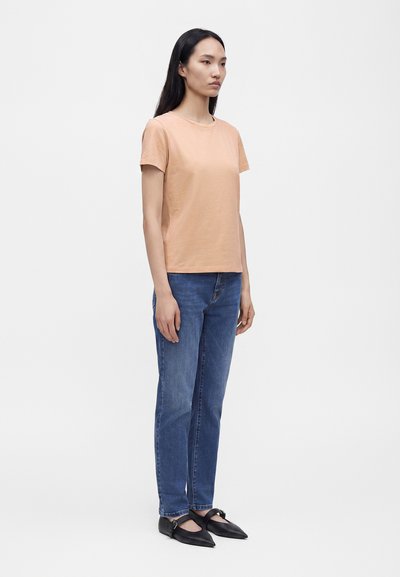 Femme aux longs cheveux foncés portant un t-shirt pêche à manches courtes, un jean bleu et des ballerines noires pointues, debout devant un fond blanc uni.