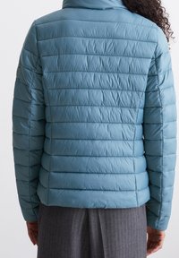 Hellblaue gepolsterte Jacke mit horizontalen Nähten, hohem Kragen und strukturierter Oberfläche. Verfügt über einen dezenten Logo-Patch am Ärmel.