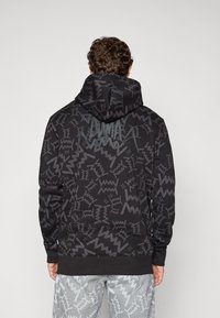 Mand iført sort hættetrøje med grå abstrakte zigzag-mønster og "PUMA"-logo på ryggen, kombineret med grå mønstrede bukser, vendt væk.