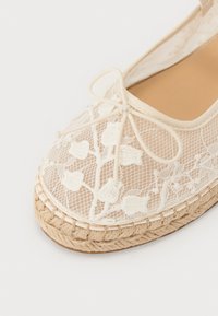 Espadrille in pizzo con tomaia in pizzo beige caratterizzata da motivi floreali, punta rotonda e suola in juta intrecciata. Accentatura a fiocco sulla parte anteriore.
