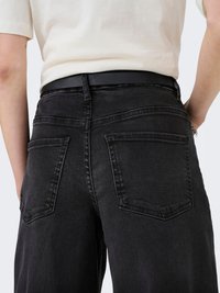Person trägt schwarze Jeans mit großen Gesäßtaschen und einem schwarzen Gürtel, kombiniert mit einem in die Hose gesteckten beigen Hemd.