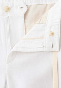 Gros plan d'un vêtement blanc avec un bouton, une fermeture éclair et une doublure intérieure partiellement visible.