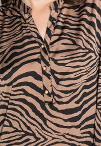 Brun och svart zebra-randig blus med v-ringning och tre guldknappar som accentuerar framsidan. Mjuk, slät tyg med en lätt lyster.