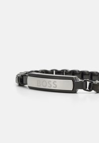 Pulsera de metal en negro y plata con una placa central ovalada grabada con "BOSS", que presenta un diseño de cadena interconectada y textura suave.