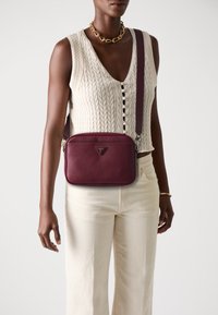 Donna che indossa un top senza maniche a maglia a trecce color crema e pantaloni, accessoriate con collana in catena dorata, bracciale e borsa a tracolla bordeaux.