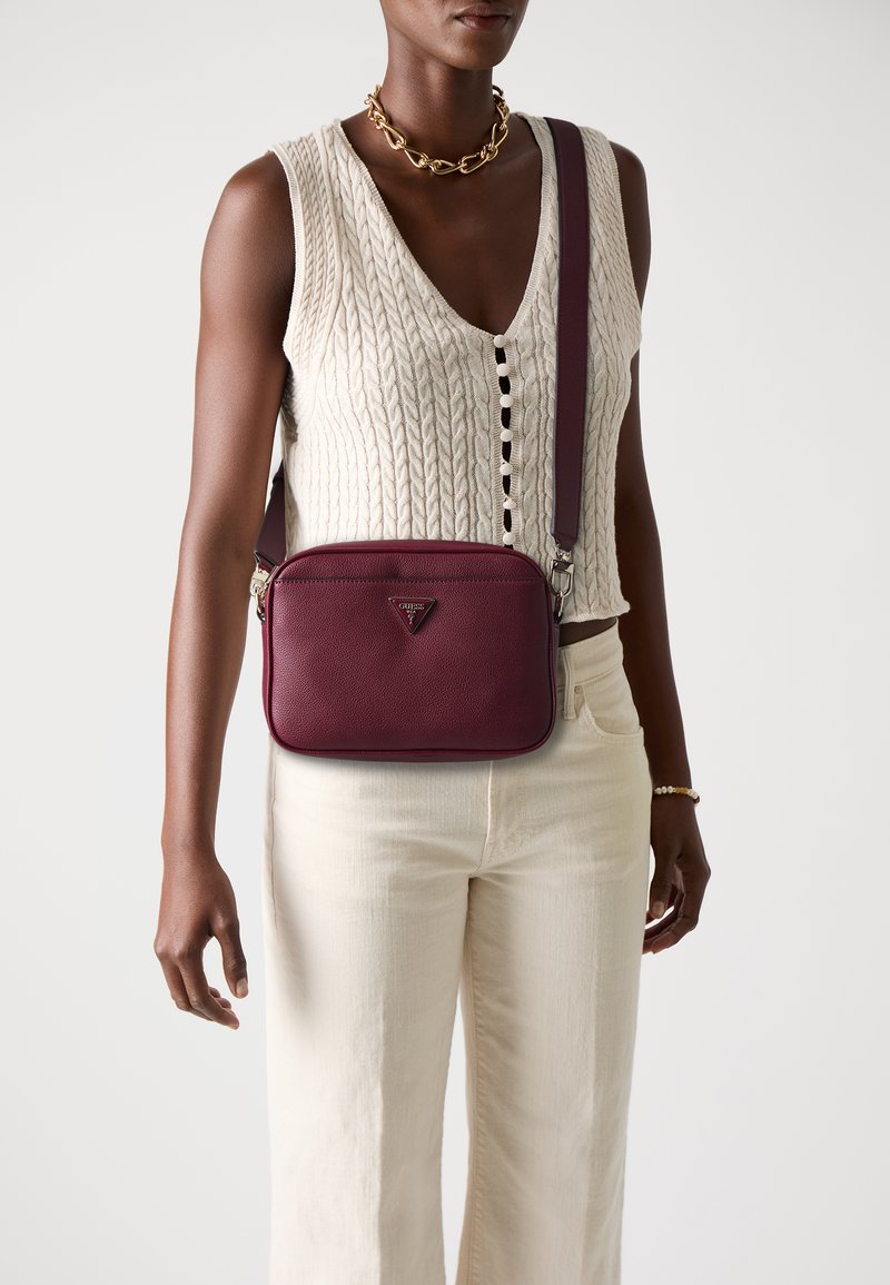Donna che indossa un top senza maniche a maglia a trecce color crema e pantaloni, accessoriate con collana in catena dorata, bracciale e borsa a tracolla bordeaux.