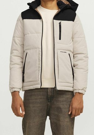 Mand iført beige og sort polstret jakke med lynlåslommer over hvid sweatshirt og mørke jeans, stående mod en ensfarvet baggrund.