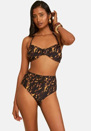 Kobieta w brązowo-czarnym wzorzystym bikini z wysokim stanem i okrągłymi klamerkami, pozująca z jedną ręką dotykającą włosów.