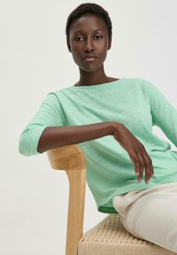 Chemise long-sleeve texturée vert clair avec un large décolleté, portée de manière décontractée sur un pantalon clair. Mannequin assis sur une chaise en bois.