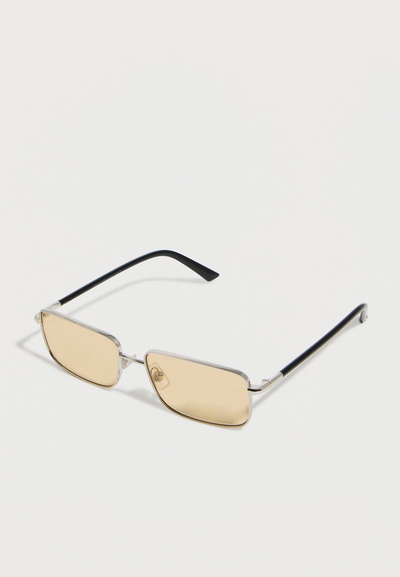 Γυαλιά ηλίου με ασημένιο πλαίσιο, μικρούς ορθογώνιους φακούς με κεχριμπαρένια απόχρωση και μαύρους βραχίονες, πάνω σε λευκό φόντο.