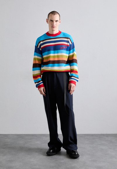 Pull en tricot color�é avec des rayures horizontales en bleu, rouge, vert et marron, associé à un pantalon noir à jambes larges et des chaussures noires épaisses.