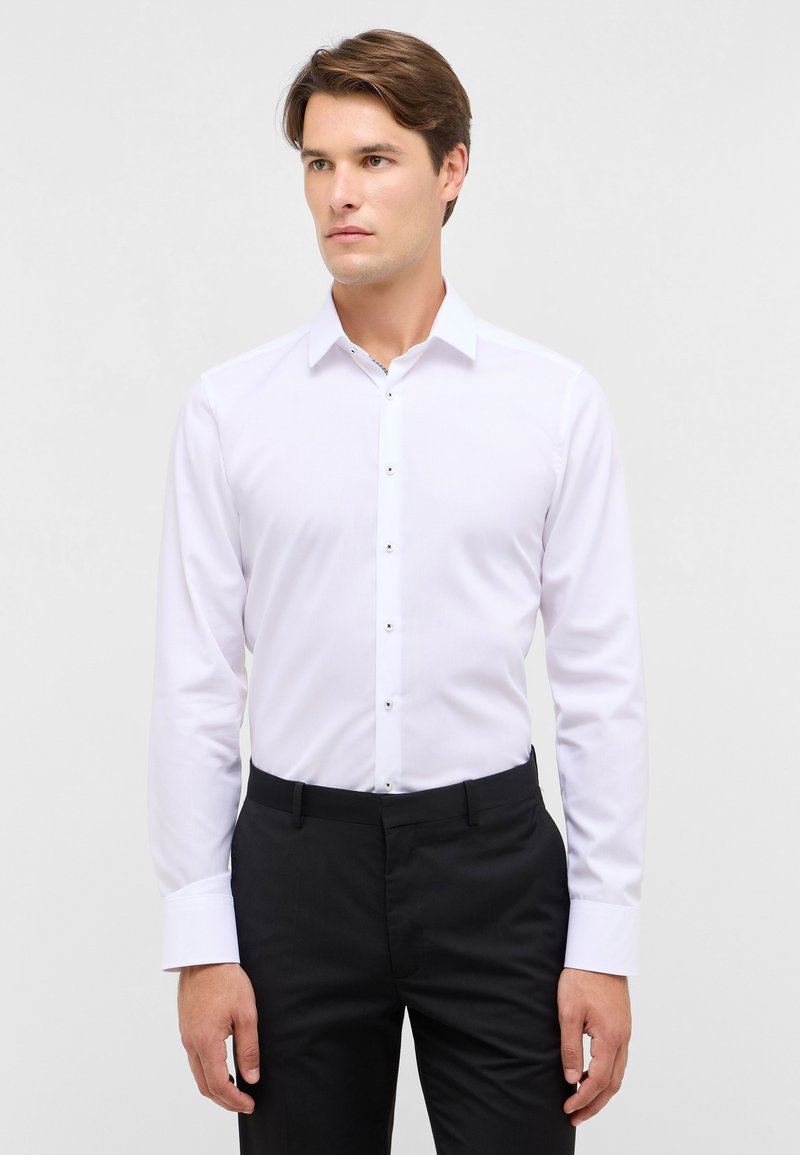 Camicia bianca a maniche lunghe con colletto a punta, caratterizzata da bottoni neri sul fronte. Tessuto liscio e vestibilità su misura.