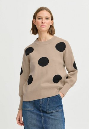 Beige Pullover mit großen schwarzen Pünktchen, Rundhalsausschnitt und gerippten Bündchen; kombiniert mit einem Jeansrock mit Taschen.