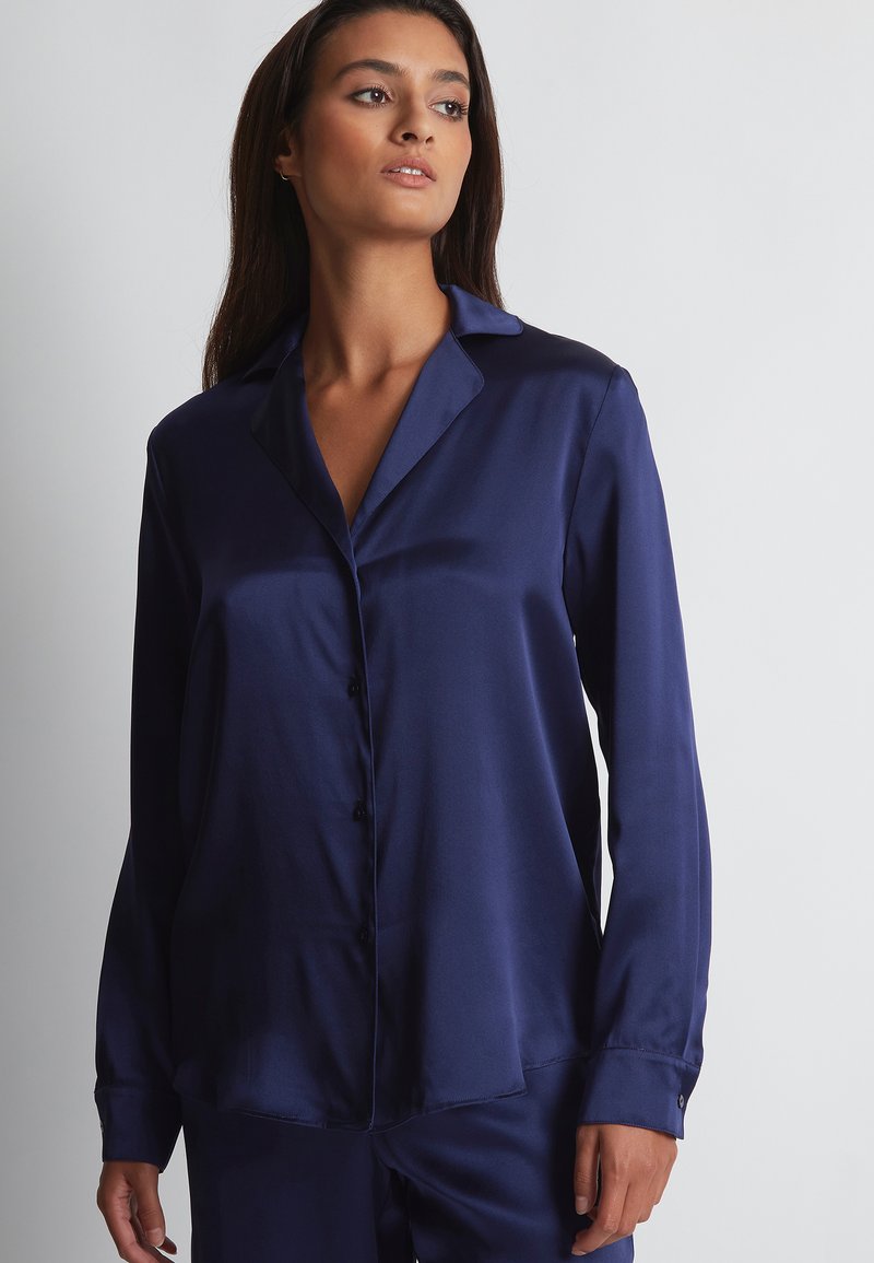 Chemise en satin bleu marine avec un col classique, des manches longues et une fermeture à boutons sur le devant. Texture lisse, coupe décontractée et subtil éclat.