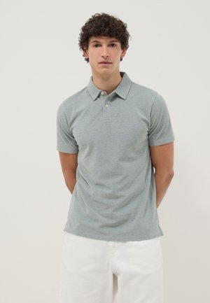 Jeune homme aux cheveux bouclés portant un polo gris clair à manches courtes et un pantalon blanc, debout, les mains dans le dos.