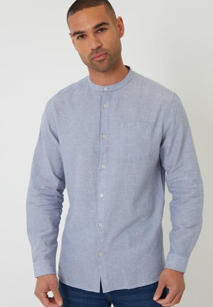 Homme portant une chemise bleu clair à boutons avec col mao, debout devant un fond blanc uni.