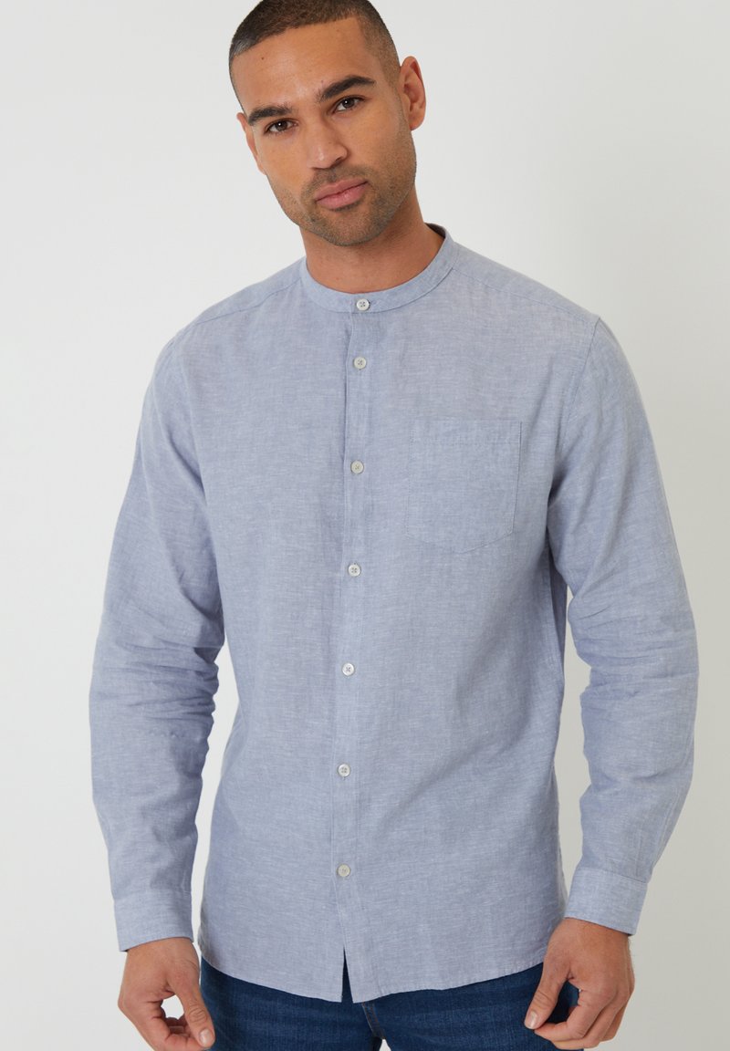 Mann, der ein hellblaues, knöpfbares Hemd mit Stehkragen trägt, steht vor einem schlichten weißen Hintergrund.