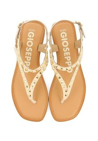 Sandalias planas beige con correas con tachuelas y una suela de goma marrón. Presentan un diseño en T con una correa en el tobillo para un ajuste seguro. Logo de la marca visible.