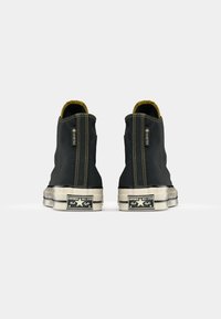 Zapatillas altas negras con acentos amarillos, fabricadas con una tela texturizada. Cuentan con suelas de goma blanca y un clásico logo de Converse All Star en el talón.