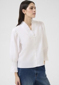 Bluză albă cu decolteu volan, închidere cu butoane și mâneci bufante cu tiv elastic. Material neted și croială lejeră. Purtată cu blugi albastre.
