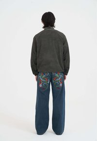 Mørkegrøn fløjlsjakke med høj krave, parret med løse blå denim jeans med indviklet flerfarvet dragebroderi på bagsiden.