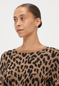 Tijgerprint trui in tan met zwarte vlekken. Zachte gebreide stof, ontwerp met ronde hals, en ribgebreide kraagdetails. Zichtbare zilveren ringoorbel.