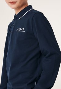 Jūras zila gara piedurkne polo krekls ar balta kontrasta apkakli, kuram ir teksturēta auduma struktūra un izšūts "BAKER by TED BAKER" logo uz krūtīm.