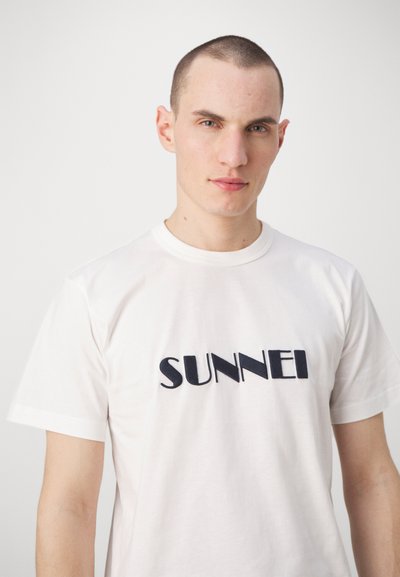 Tricou alb din bumbac cu mâneci scurte, având cuvântul "SUNNEI" brodat în litere albastre închise pe piept.