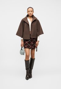 Γυναίκα που φοράει καφέ κάπα-τζάκετ, λευκό τοπ, καρώ μίνι φούστα, μαύρες μπότες μέχρι το γόνατο και κρατάει μια μικρή ανοιχτό μπλε τσάντα.