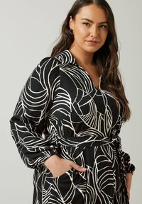 Zwarte jurk met witte abstracte bladerenpatronen, kraag, lange mouwen, ceintuur in de taille en zakken. Textuurstof met een vloeiend ontwerp.