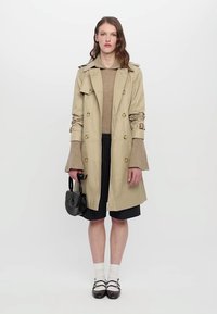 Khaki trench-coat, jossa on kaksirivinen malli, vyölliset hihat, yhdistetty ruskeaan neuleeseen, mustiin raidallisiin shortseihin ja mustaan käsilaukkuun.