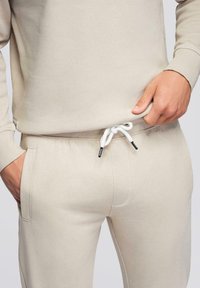 Homme portant un pantalon de survêtement beige avec un cordon blanc et un sweat-shirt assorti, ajustant l'ourlet du sweat-shirt d'une main, l'autre main dans la poche.