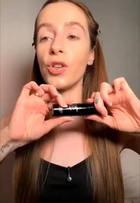 Femme aux longs cheveux bruns tenant un produit cosmétique cylindrique noir près de son visage, parlant devant un fond uni.