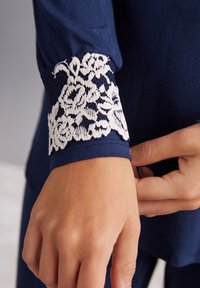 Marineblauwe long-sleeve top met een witte kantafwerking bij de manchetten. De stof is glad met een lichte glans. Het kant heeft een bloemmotief.
