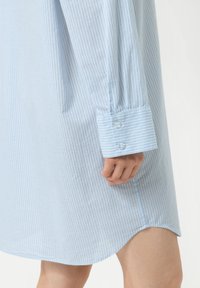 Robe chemise rayée bleu clair, à manches longues et poignets boutonnés. Le tissu semble lisse avec une texture subtile. L'ourlet est légèrement courbé.