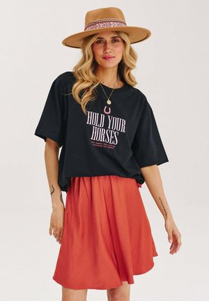 Czarny t-shirt z napisem "HOLD YOUR HORSES", zestawiony z zwiewną pomarańczową spódnicą i beżowym kapeluszem z wzorzystą bandą. Miękkie tekstury materiałów.