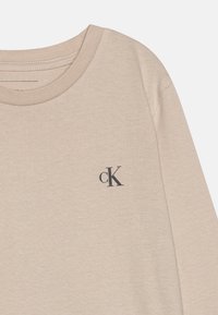Beige ilgi piedurkņu krekls, kas izgatavots no mīkstas kokvilnas, ar apaļu kakla izgriezumu un mazu melnu "cK" logotipu uz krūtīm. Gluda tekstūra, ikdienišķs dizains.