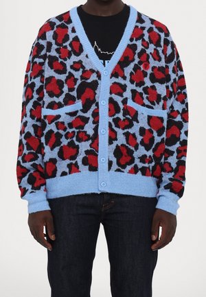 Mann trägt eine blaue, geknöpfte Strickjacke mit roten und schwarzen Leopardenflecken, zwei vorderen Taschen, schwarzem Hemd und dunkler Jeans.