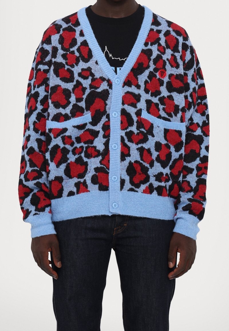 Homme portant un cardigan bleu boutonné avec des taches léopard rouges et noires, deux poches avant, une chemise noire et un jean foncé.