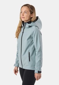 Femme aux cheveux longs et blonds, portant une veste à capuche bleu clair avec une fermeture éclair noire et des leggings noirs, debout devant un fond uni.