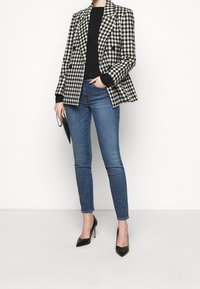 Blazer de cuadros vichy en blanco y negro de doble pecho sobre un suéter negro, combinado con jeans skinny azules y zapatos de tacón alto negros.