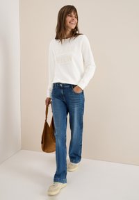 Femme souriante marchant, portant un sweatshirt blanc avec un texte, un jean bleu, des baskets beiges, tenant un sac en daim marron sur un fond neutre.