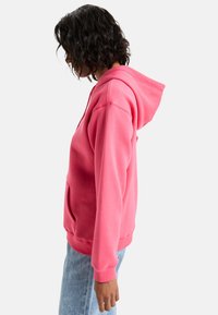 Sudadera rosa hecha de una tela suave, con un bolsillo amplio, capucha con cordón y puños acanalados. Llevada sobre unos vaqueros de color azul claro.